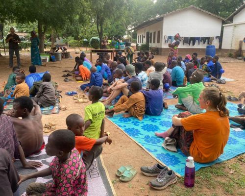 Elizabeth sits amongst a crowd of local children listening to a speaker. 