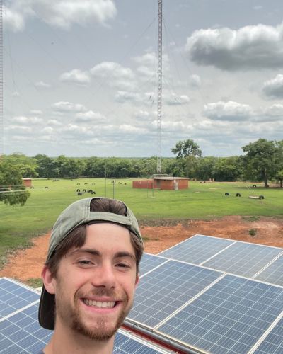 Luke smiles in the foreground with the transmitter and transmitter field behind him in the distance. 