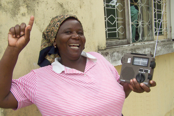 An African lady holding a TWR radio, smiling while pointing up.