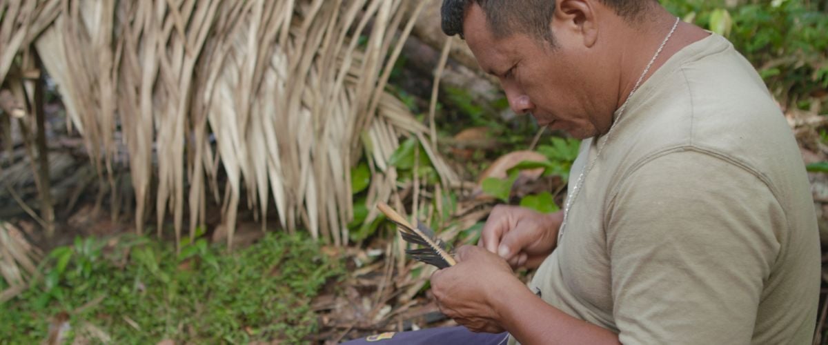 An indigenous Wayana man sits on the ground crafting an arrow. 