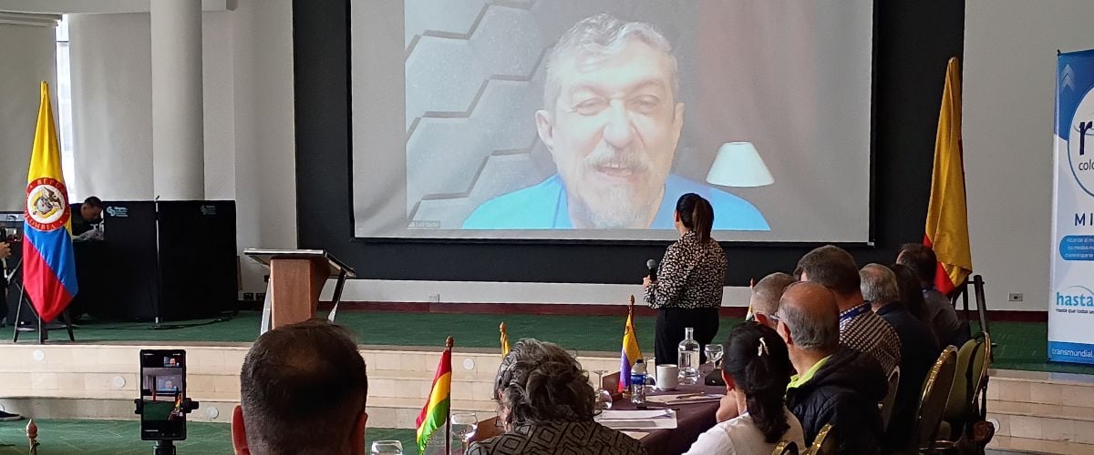 Luiz Sayão speaks via a video link during the Ibero-Latin America Partners Conference. Conference attendees sit in the foreground, watching the screen with Lui's face projected in the background.
