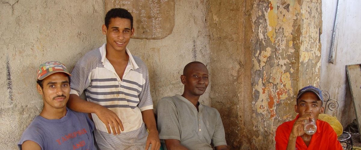 Four Cuban men sit and stand against a wall, smiling and looking at the camera.