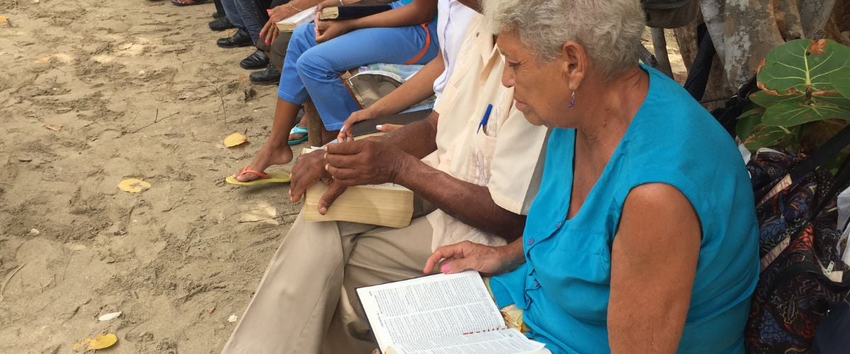 An elderly woman in Cuba sits in an outdoor church, Bible open across her lap. 
