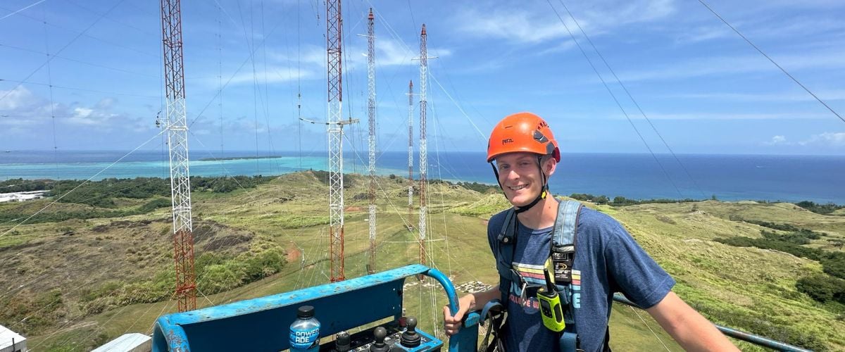 Brenic Beggs stands on a lift in the TWR antenna field on Guam. The towers stand in the background with the ocean view behind them. 