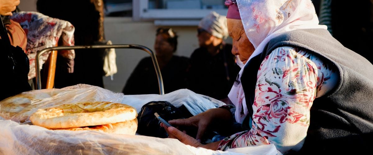 A Karakalpak woman leans over her food stand, looking down at a cell phone.