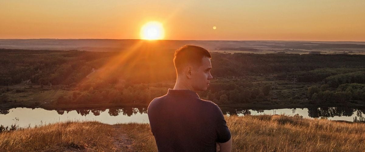 Tatar man stands with his back to the camera, looking right, as the sun sets in the distance. 