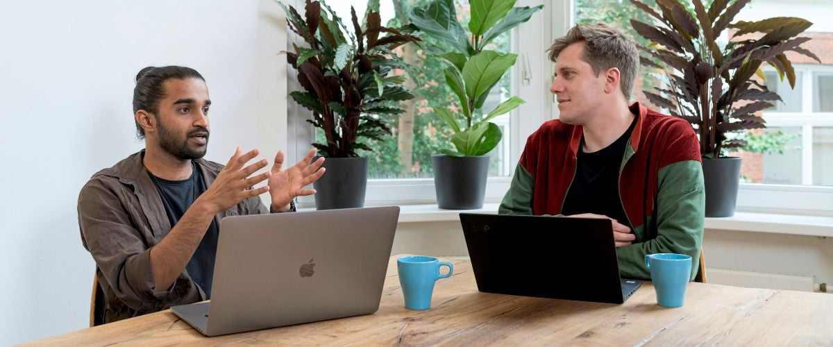 Two men discuss a project with their laptops open on a table. 