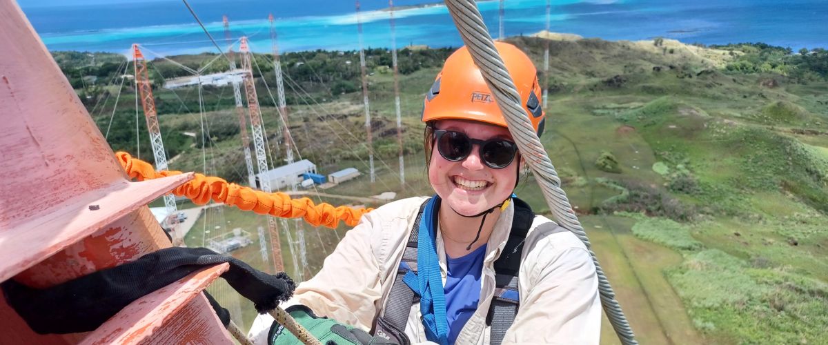 Emma Fanning smiles from the top of a TWR radio tower on Guam, the island and other towers visible in the background. 