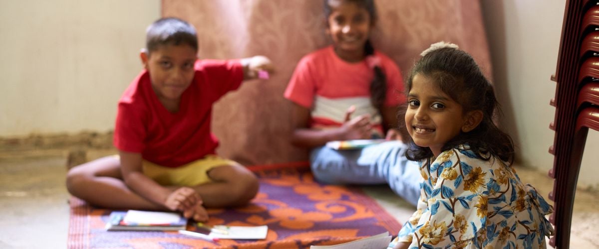 A young girl from Sri Lanka looks at the camera, smiling, while sitting on the floor before two other children.