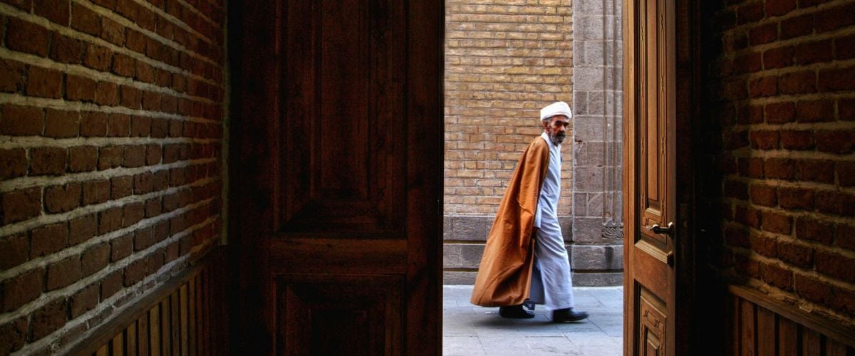 A man in traditional clothing looks in as he walks past a darkened doorway in Iran. 