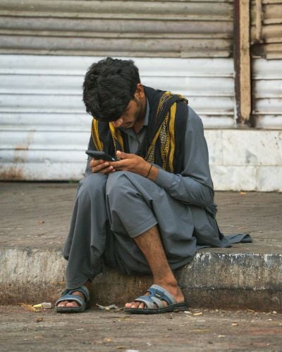 A young Pakistani man looks down at his phone while sitting on a curb at the side of a road.
