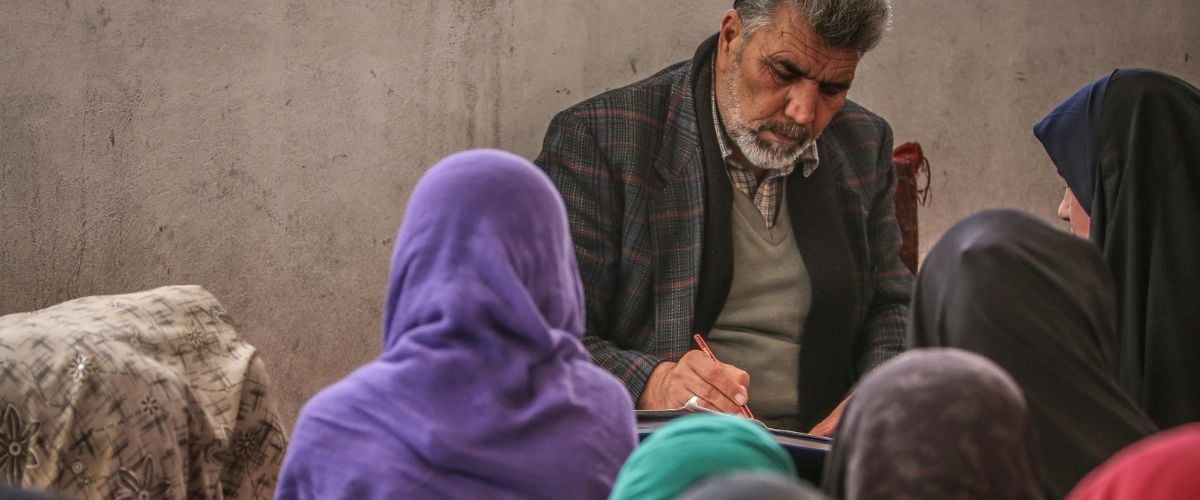 A man stands before a class of women, looking down at a notebook and writing.